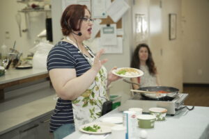 A person with short hair and glasses giving a live cooking demonstration with a hotplate and frying pan.