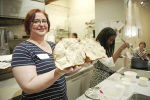 A person close to camera holds up a loaf of bread that has been ripped open, while another person in the background cooks.