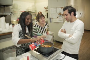 A person with long black hair cooking at a hotplate while two other people watch.