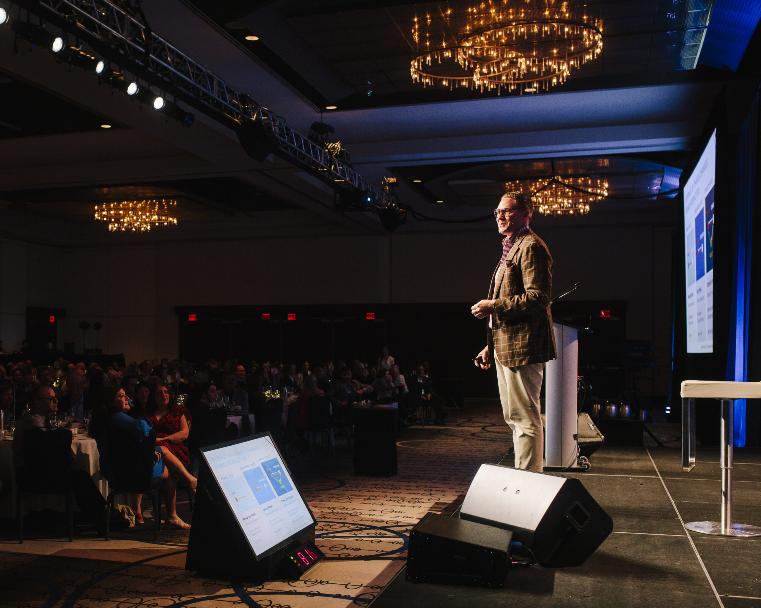 A person on stage looking out over a dark room crowded with people seated around tables.