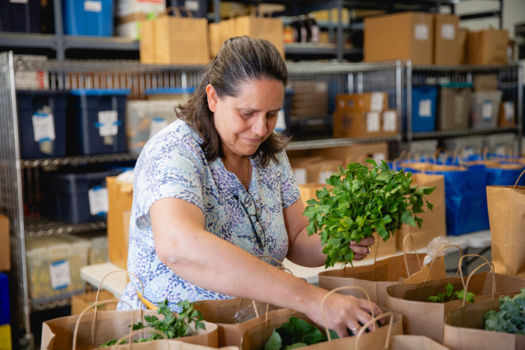 A person with medium length hair puts leafy green vegetables into multiple brown grocery bags.