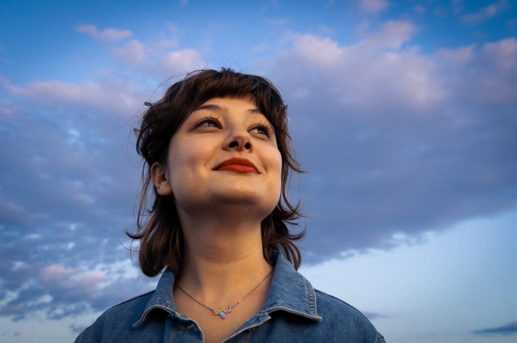 Photograph of a person with dark, shoulder-length hair wearing a denim jacket and smiling towards the sun, taken in the sweet pastel colors of the sunset.