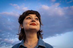 Photograph of a person with dark, shoulder-length hair wearing a denim jacket and smiling towards the sun, taken in the sweet pastel colors of the sunset.