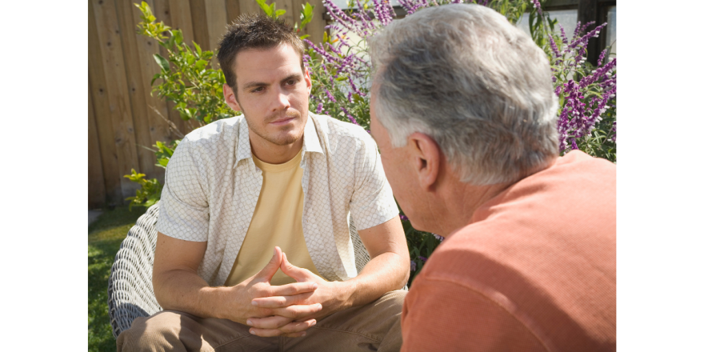 Two people sitting outside in conversation. A young person with short hair listens seriously to an older person facing away from the camera.