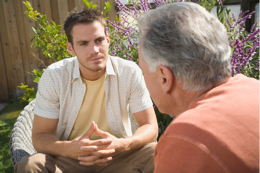 Two people sitting outside in conversation. A young person with short hair listens seriously to an older person facing away from the camera.